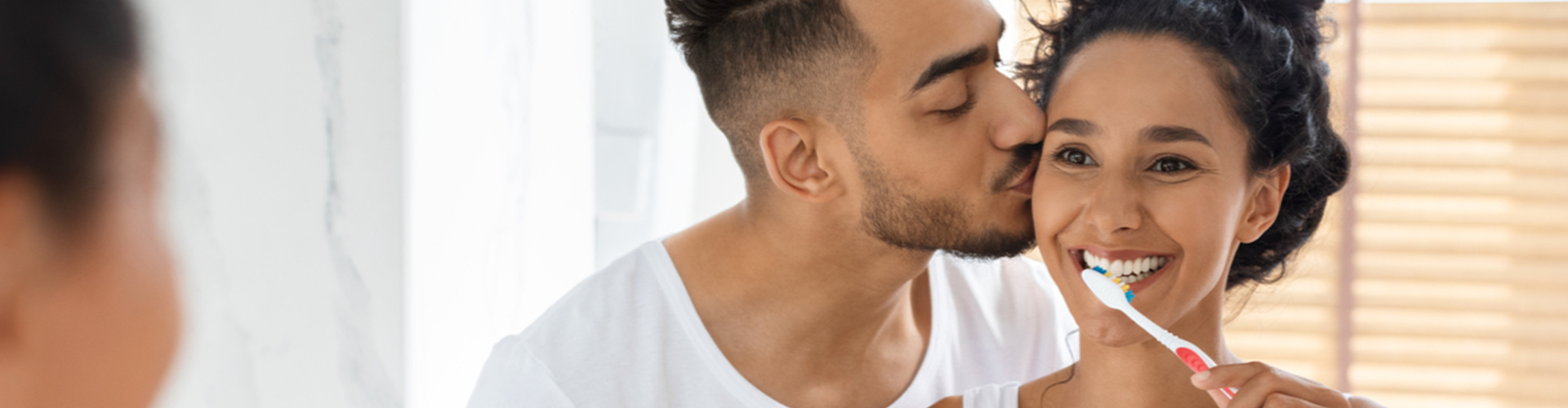 woman brushing her teeth and partner kissing her on the cheek