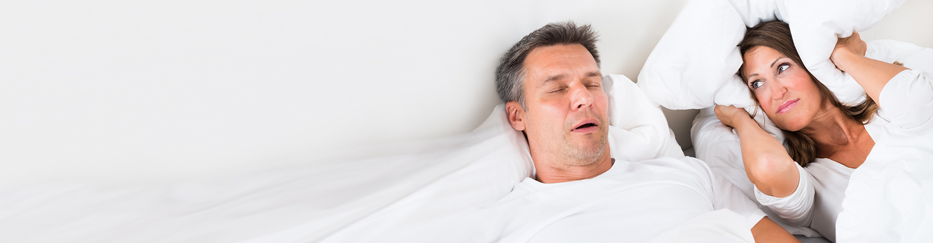 man sleeping on back with mouth open while woman covers own ears with pillow